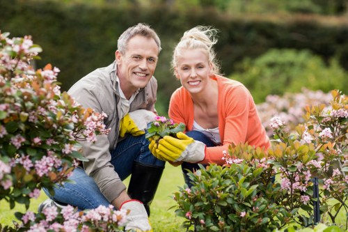 A middle-aged man and a young woman are working together in a well-maintained garden, surrounded by flowering bushes with pink and white blossoms. They are kneeling on the grass, which is lush, green, and evenly mowed, and they are smiling at the camera. The man is wearing a light grey shirt and jeans, holding gardening gloves, while the woman is in an orange jumper and white shirt, also wearing gardening gloves. The background shows a hedge and trees, with soft natural light indicating a clear, sunny day, typical of outdoor gardening work in Hoxton. Their activity suggests they are engaging in garden maintenance or planting, and the setting features a tidy, landscaped outdoor space with healthy plants and a neat lawn, highlighting professional gardening services offered by Lawn Mowing Hoxton for residential outdoor spaces.